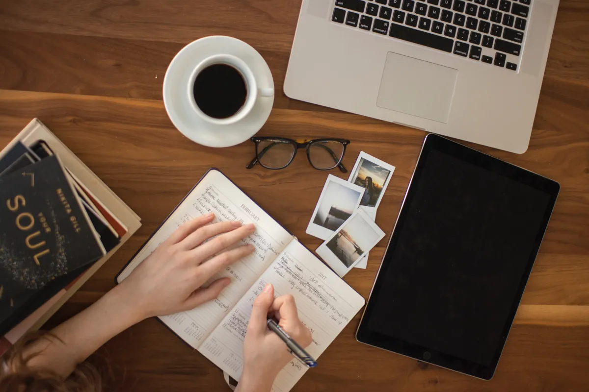 Wooden desk with a laptop, a tablet, a cup of coffee, glasses, books and someone writing in a notebook.
